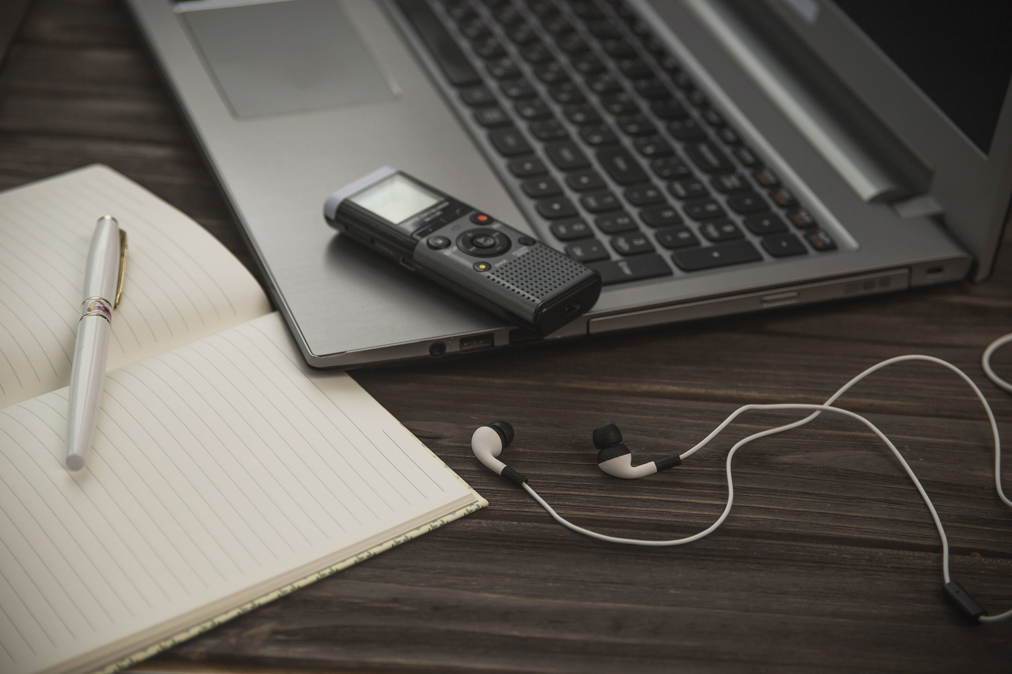 A notebook, pen, laptop, voice recorder and headphones arranged in a stock photo demonstrating the tools of a journalist.