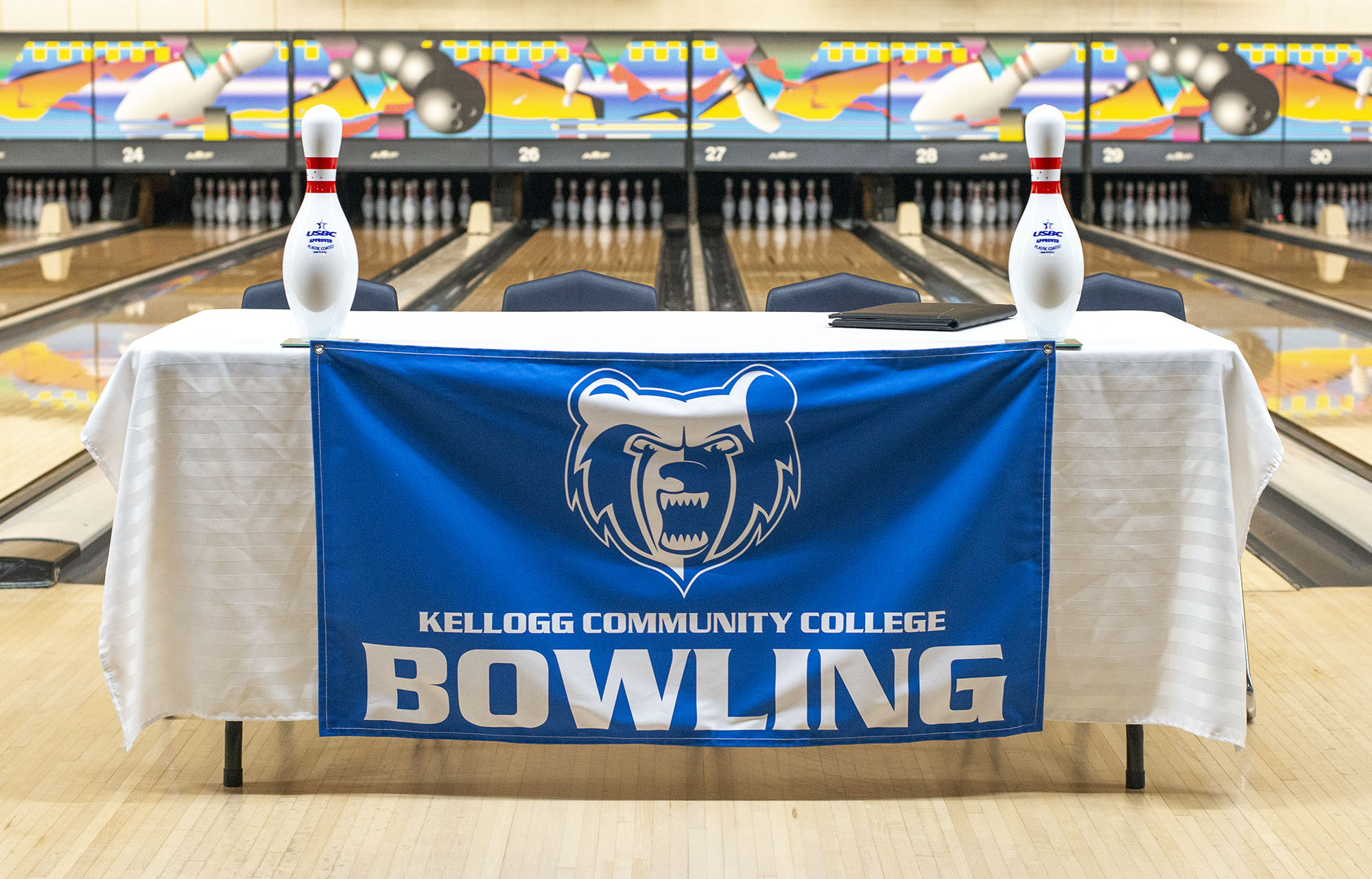 A table in a bowling alley covered with a cloth featuring the KCC logo and the words "Kellogg Community College Bowling."