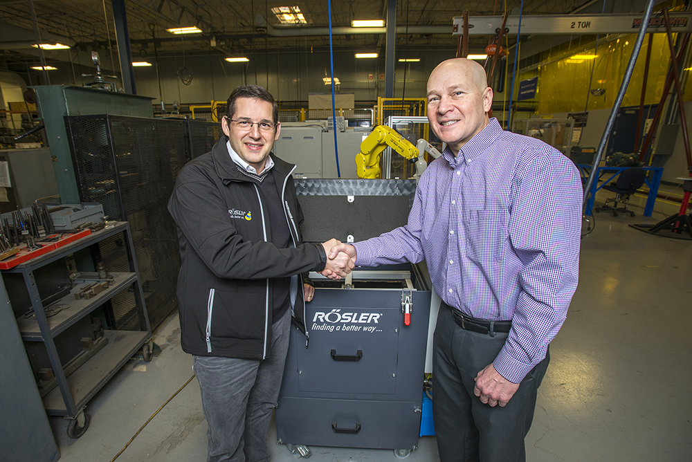 Rosler CEO Bernhard Kerschbaum, left, and RMTC Director Tom Longman shake hands in front of a new vibratory tub finishing machine donated by Rosler.