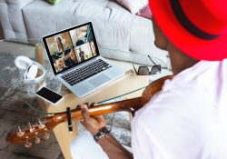 A man playing guitar watches a laptop screen where four other people are singing and/or playing instruments.