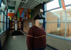 A student wears a virtual reality headset while standing in a hallway on campus.
