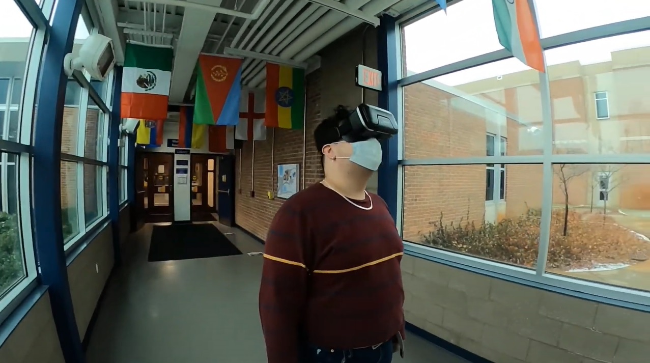 A student wears a virtual reality headset while standing in a hallway on campus.