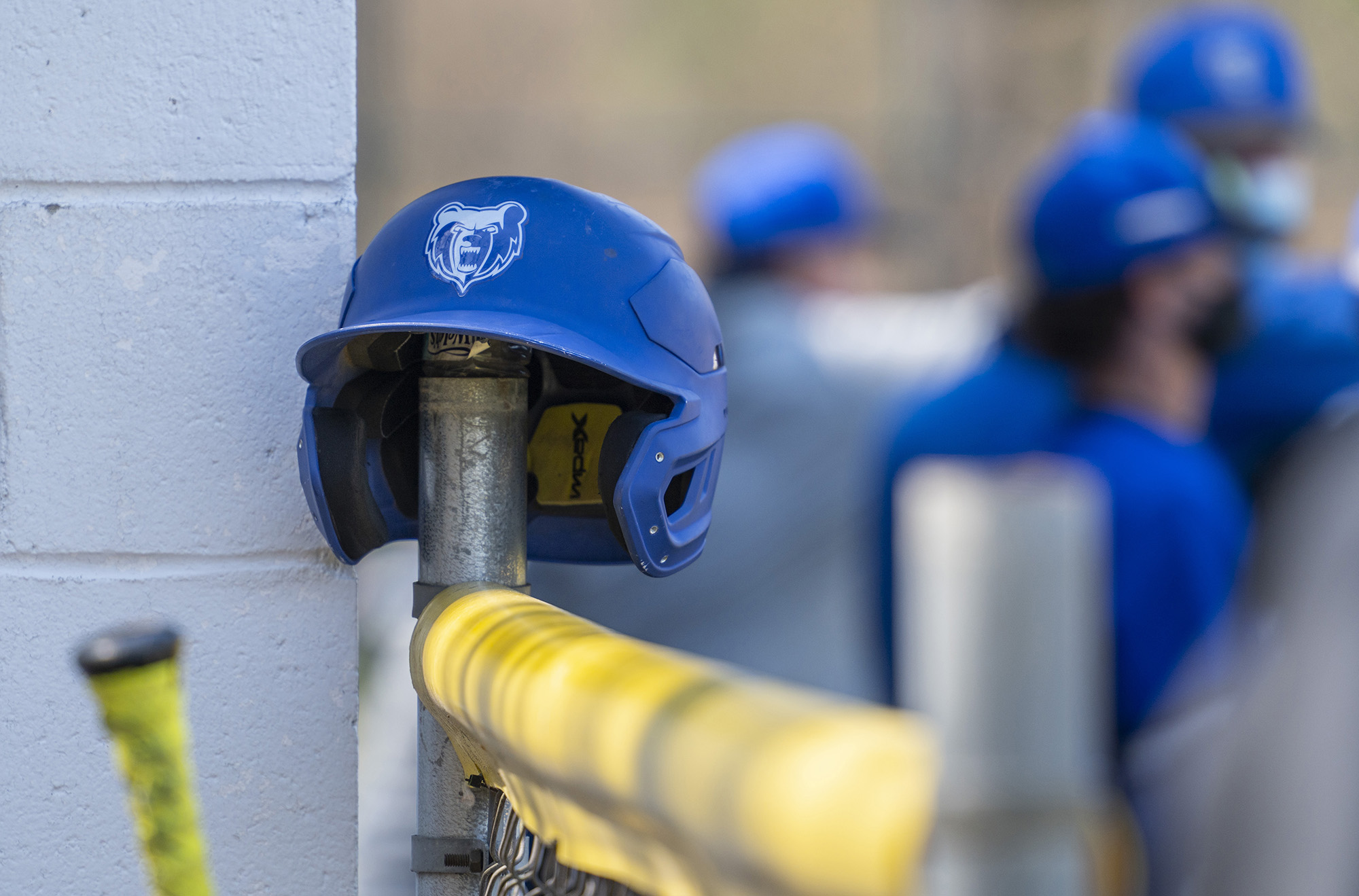 A batting helmet with the KCC Bruin head logo on it sits on a fence during a game.