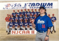 A KCC baseball player in a new jersey poses in front of a photo of the 1999 KCC baseball team.