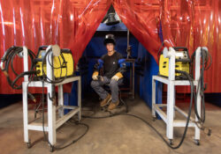 Aria dressed in welding gear and posing for a photo in a booth in the Welding Lab.