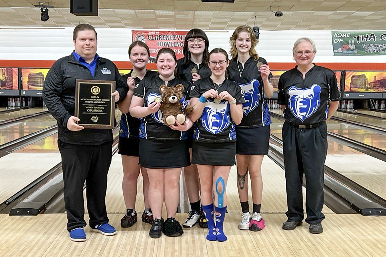 The women's bowling team and coaches pose with awards in a bowling alley.