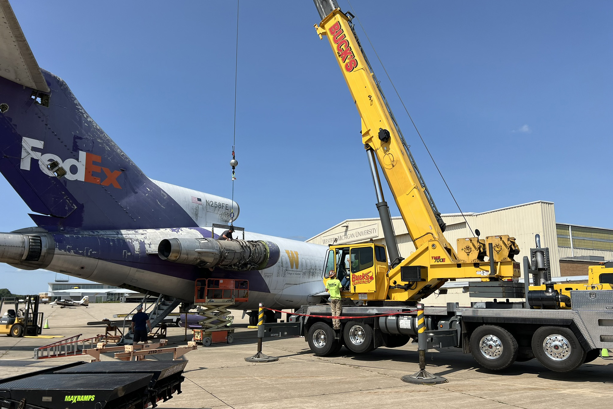 A crane removes a jet engine from the side of an airplane.