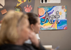 An artistic picture with text on it that says "art" hangs on the wall of a classroom as students in an art class listen to their instructor.