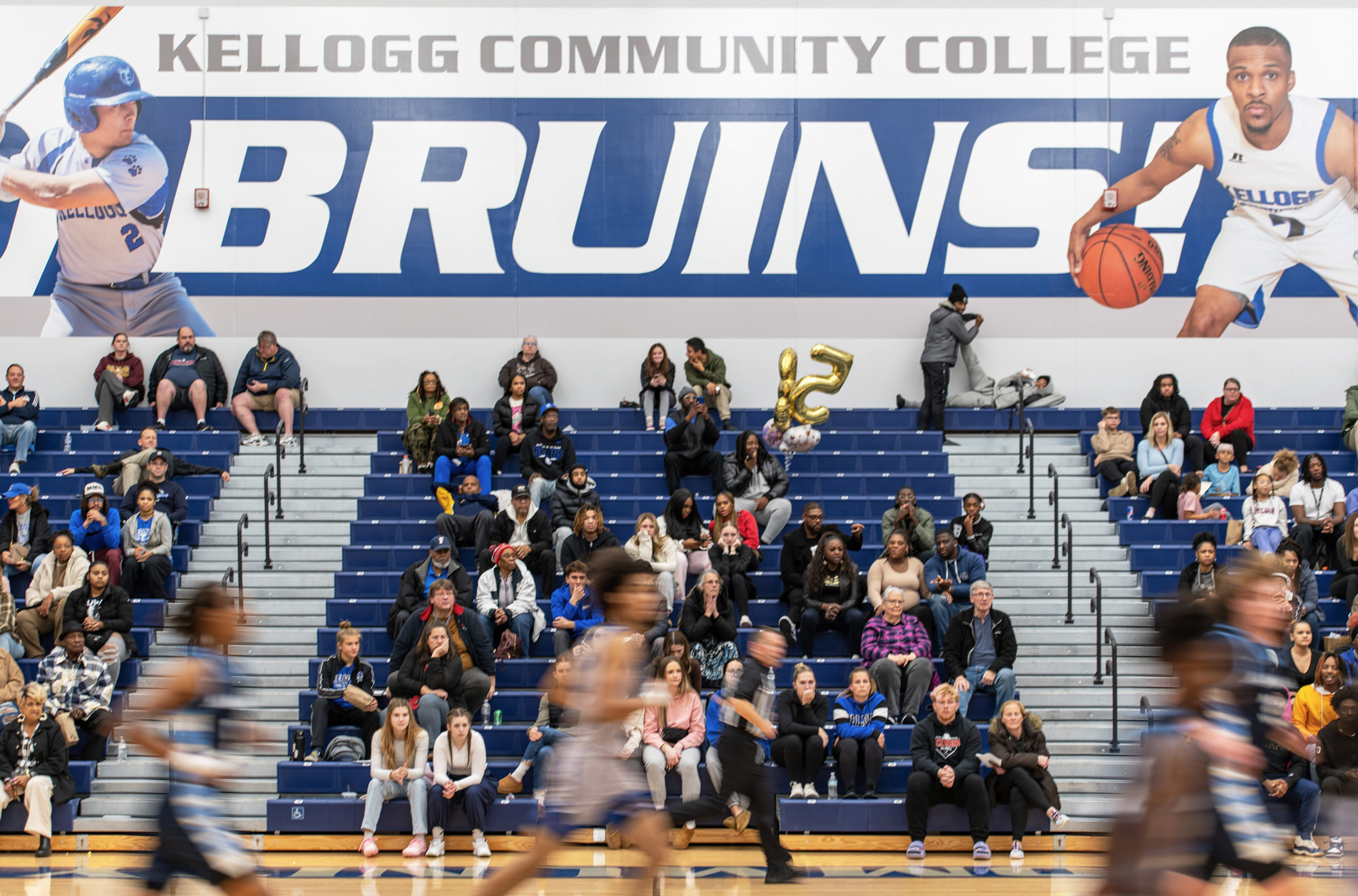 A banner reading "Kellogg Community College Bruins" hangs on the gym wall over fans in the stands during a KCC home basketball game.