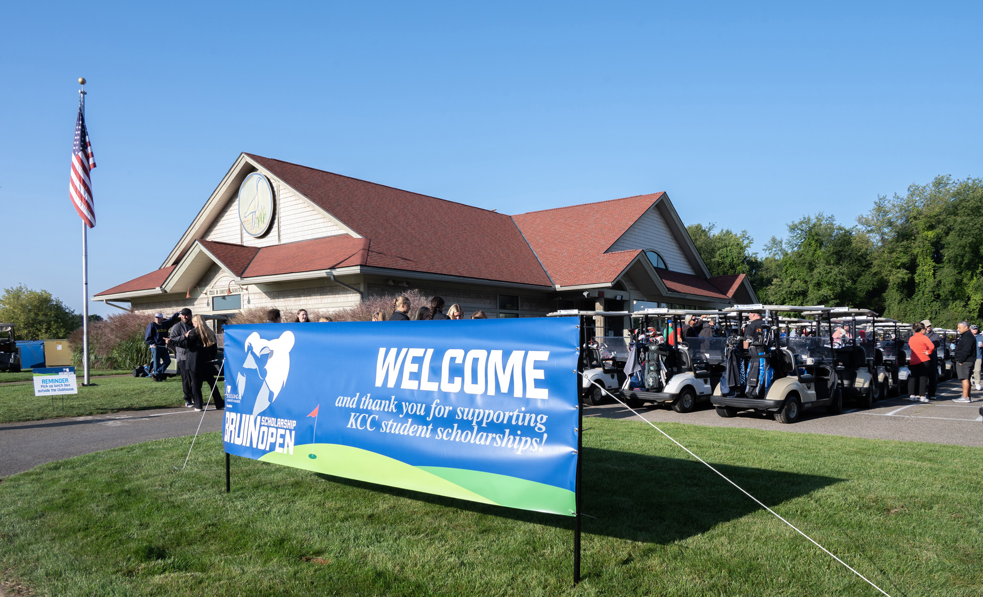 A banner that reads, "Welcome and thank you for supporting KCC student scholarships!" stands in front of a golf course clubhouse and lines of golf carts at the Bruin Scholarship Open.