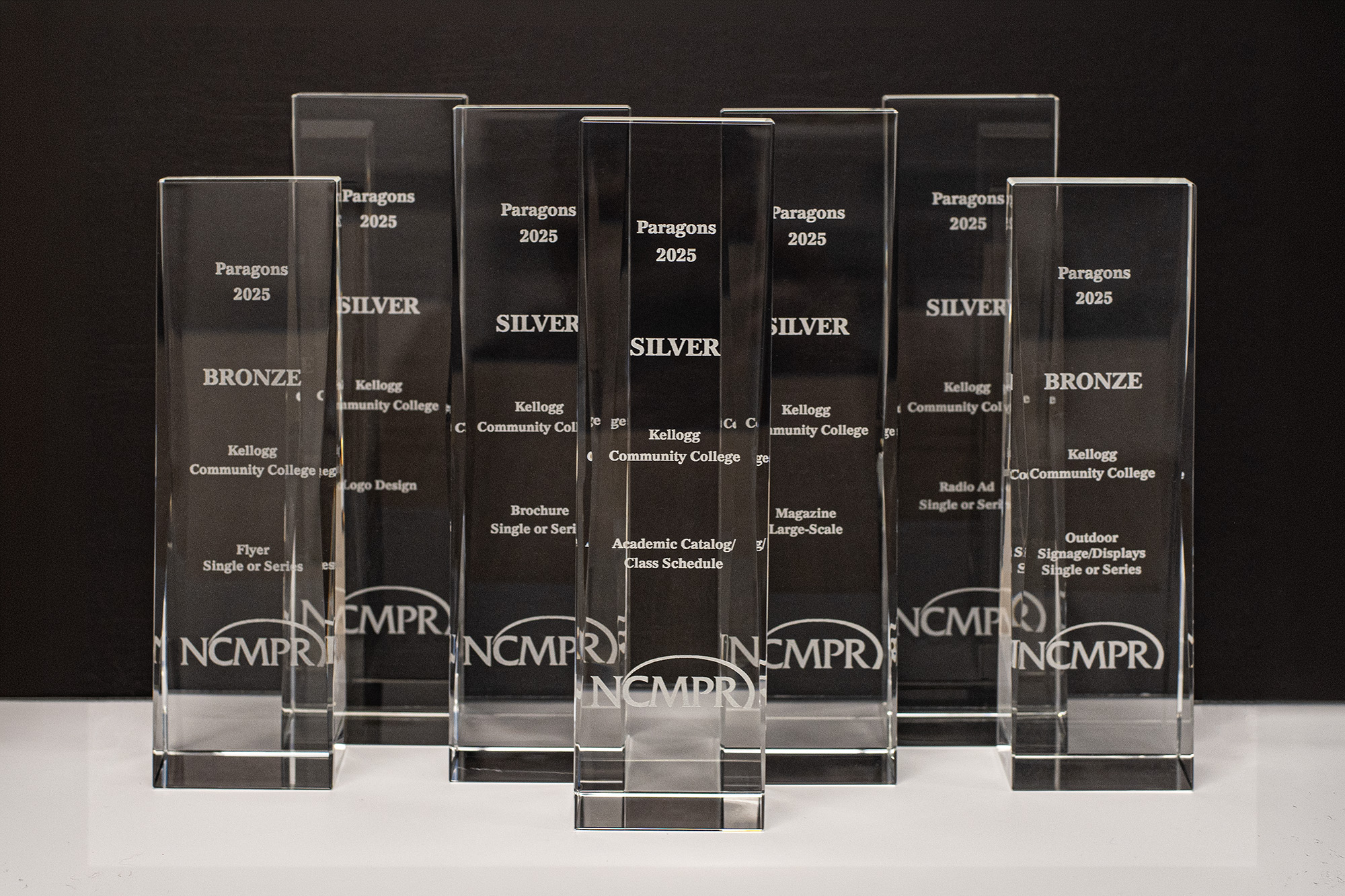 A photo of seven glass statue/trophy awards engraved with Kellogg Community College’s name, the NCMPR logo and SILVER or BRONZE designations.