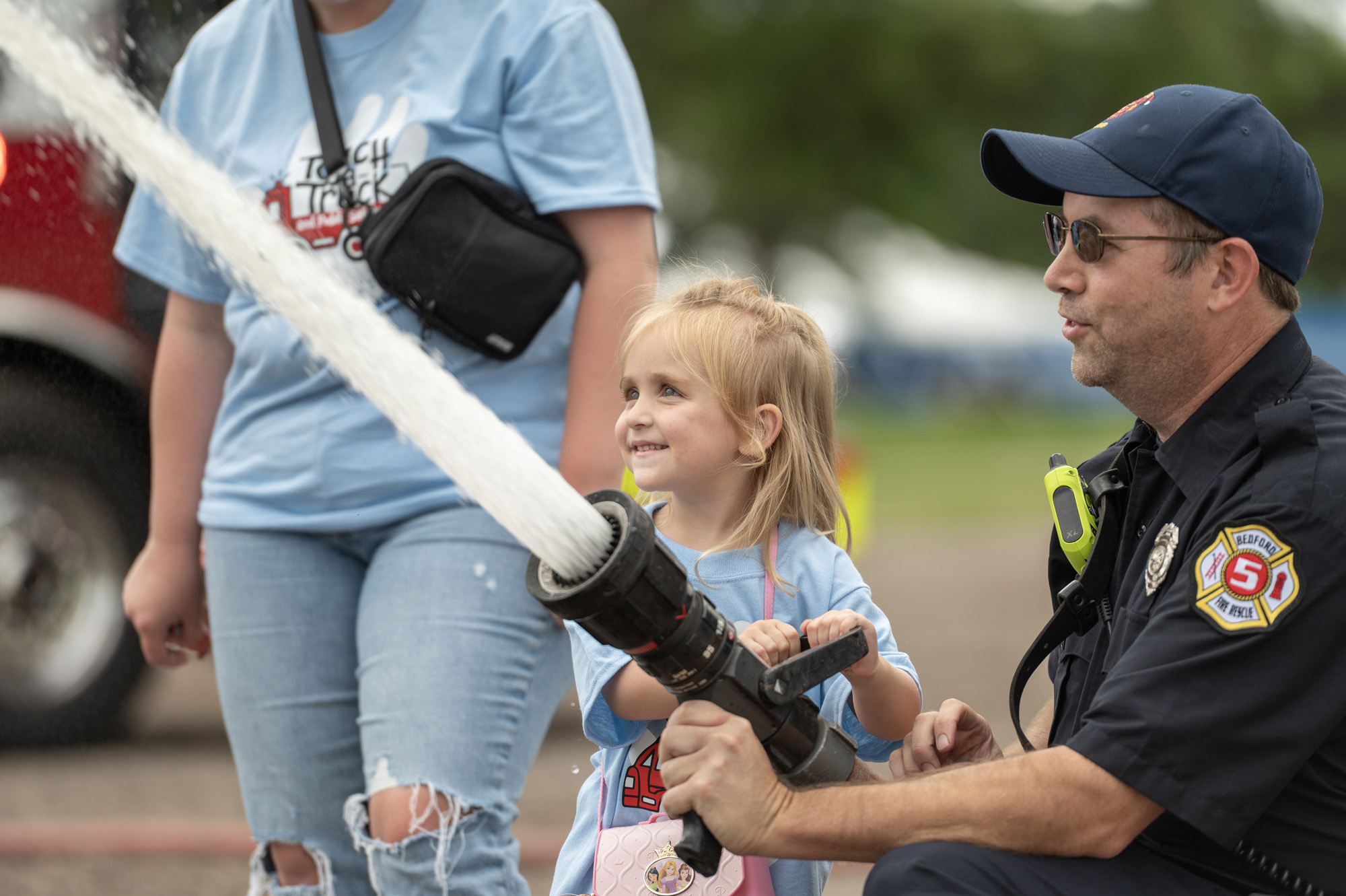A public safety official assists a young girl with using a firehose during a Touch a Truck and Public Safety Day event on campus.